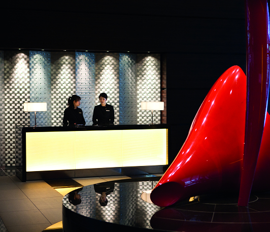 Two female front desk workers talk as they stand behind the reception desk facing a large abstract red statue